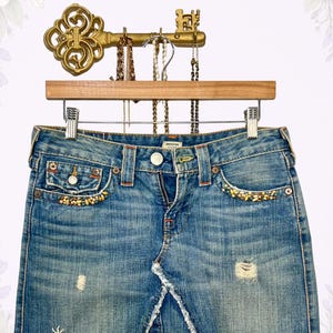 May include: Light blue denim skirt with distressed details, featuring a frayed hem and decorative beading along the pockets. The skirt is displayed on a wooden hanger with a vintage key-shaped jewelry holder above.