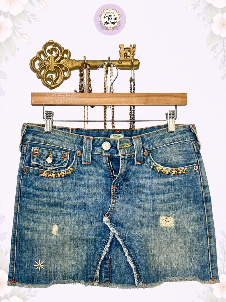 May include: Light blue denim skirt with distressed details, featuring a frayed hem and decorative beading along the pockets. The skirt is displayed on a wooden hanger with a vintage key-shaped jewelry holder above.