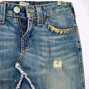 May include: Blue denim skirt with a distressed look, featuring a frayed hem and a small ripped area. The front pocket is embellished with colorful beads. The skirt has a button and zipper closure, with orange stitching details.