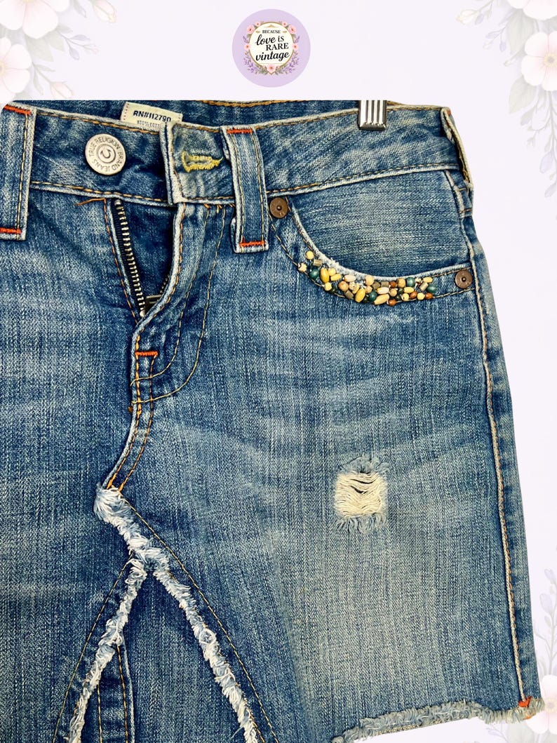 May include: Blue denim skirt with a distressed look, featuring a frayed hem and a small ripped area. The front pocket is embellished with colorful beads. The skirt has a button and zipper closure, with orange stitching details.