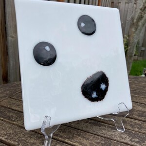 May include: White square glass plate with three black circles and a black and white abstract design. The plate is on a clear acrylic stand.