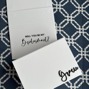 May include: White card with the text "Will you be my Bridesmaid?" and a white envelope with the name "Grace" written in black cursive.