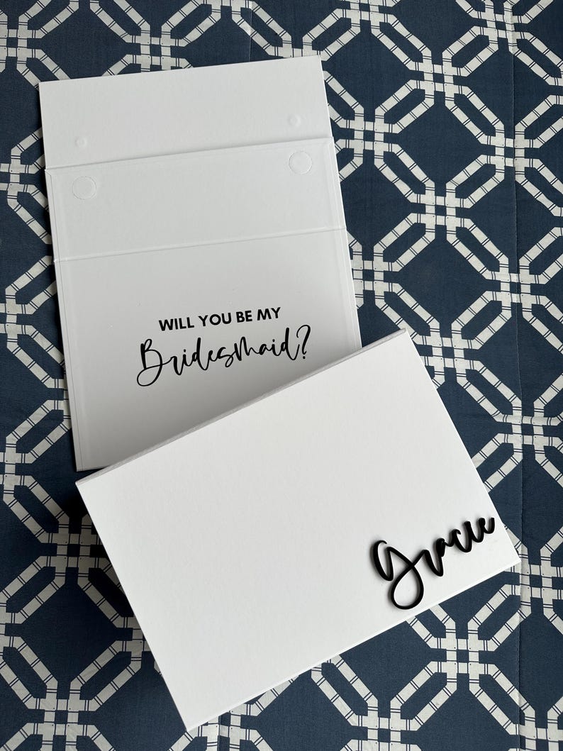 May include: White card with the text "Will you be my Bridesmaid?" and a white envelope with the name "Grace" written in black cursive.