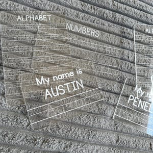 May include: Three clear acrylic dry-erase boards with white lines and text. The boards are for practicing writing letters, numbers, and names. The text on the boards reads "Alphabet", "Numbers", "My name is Austin", and "My name is Penelope".