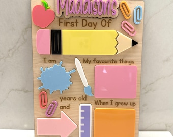 May include: A wooden sign with the text "Maddions's First Day Of" and spaces for writing about a child's first day of school. The sign has a pink apple, a yellow pencil, a blue paint splatter, a white paintbrush, a pink rectangle, a ruler, and an orange rectangle.