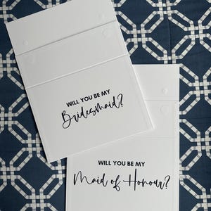 May include: Two white cards with black text. The top card says "Will you be my Bridesmaid?" and the bottom card says "Will you be my Maid of Honour?"