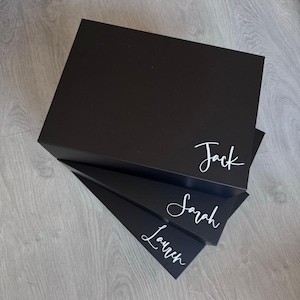 May include: Three black gift boxes stacked on top of each other. The top box has the name "Jack" written in white cursive lettering. The middle box has the name "Sarah" written in white cursive lettering. The bottom box has the name "Lauren" written in white cursive lettering.