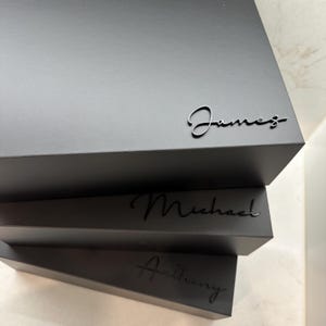 May include: Three black gift boxes stacked on top of each other. The top box has the name "James" written on it in a cursive font. The middle box has the name "Michael" written on it in a cursive font. The bottom box has the name "Anthony" written on it in a cursive font.