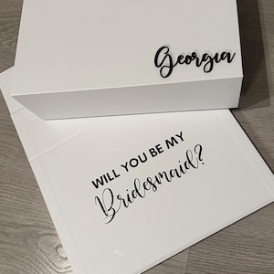 May include: White gift box with a black inscription that reads "Georgia". The box is open to reveal a white card with the text "Will you be my bridesmaid?" in black lettering.