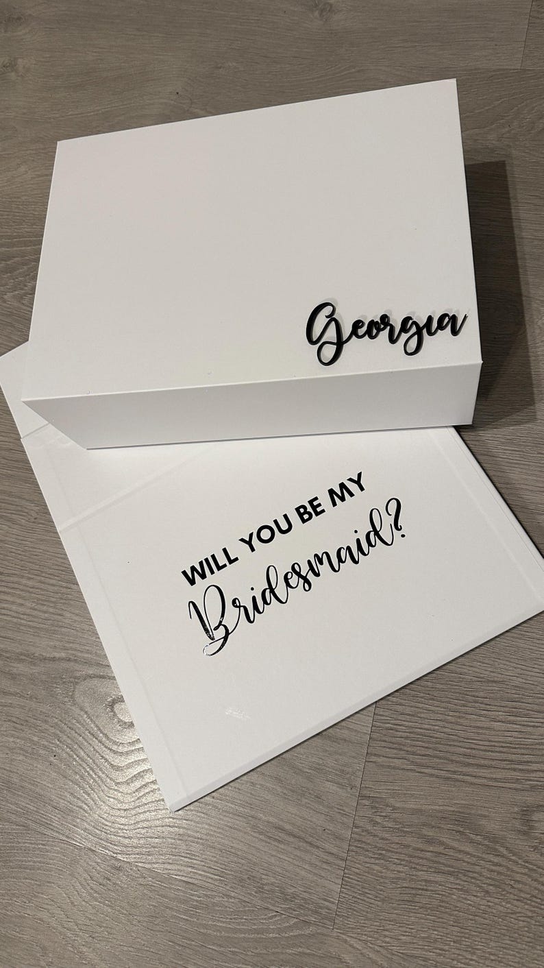 May include: White gift box with a black inscription that reads "Georgia". The box is open to reveal a white card with the text "Will you be my bridesmaid?" in black lettering.