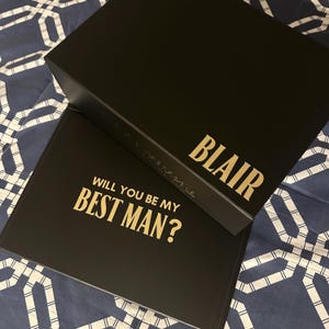 May include: Two black boxes with gold lettering. The top box has the name "BLAIR" on it. The bottom box has the text "WILL YOU BE MY BEST MAN?" on it.