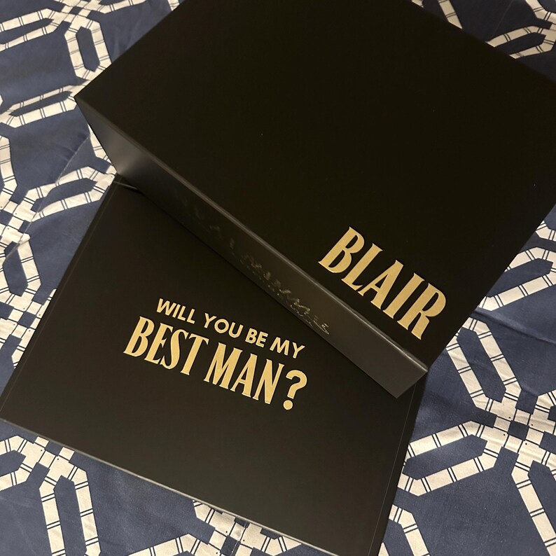 May include: Two black boxes with gold lettering. The top box has the name "BLAIR" on it. The bottom box has the text "WILL YOU BE MY BEST MAN?" on it.
