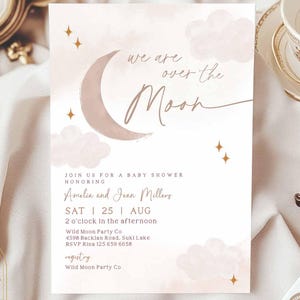 以下が含まれることがあります： 三日月と雲が描かれたピンクとゴールドのベビーシャワーの招待状。テキストには「We are over the moon」とあり、日付、時間、場所などのイベントの詳細が記載されています。