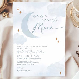 May include: A baby shower invitation featuring a blue watercolour moon and clouds. The text reads "We are over the moon" and includes details for the event, such as the date, time, and location.