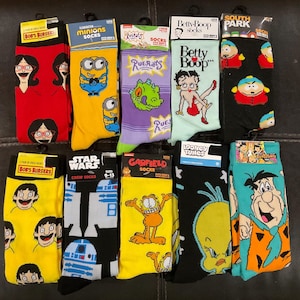 May include: Eleven pairs of crew socks featuring various cartoon characters.  The socks are in yellow, red, purple, black, teal, and light green.  Character themes include Bob's Burgers, Minions, Rugrats, Betty Boop, South Park, Star Wars, Garfield, and The Flintstones.  Sock packaging displays character names and 'socks' or 'crew socks'.