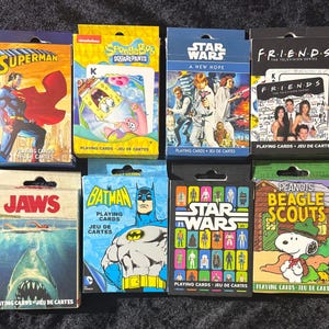 May include: A collection of playing card decks featuring various themes. The decks include Superman, SpongeBob SquarePants, Star Wars, Friends, Jaws, Batman, and Peanuts Beagle Scouts. Each deck has a unique design and is packaged in a cardboard box.
