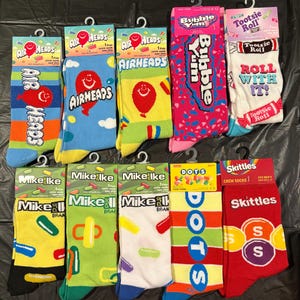 May include: A collection of colorful crew socks featuring various candy brands. Designs include Airheads, Bubble Yum, Tootsie Roll, Mike and Ike, Dots, and Skittles. Each pair is displayed with its original packaging.