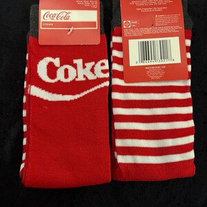 May include: Pair of red and white crew socks with the Coca-Cola logo. One sock features the word "Coke" in white, while the other has red and white stripes. The socks have a gray cuff and a red tag.