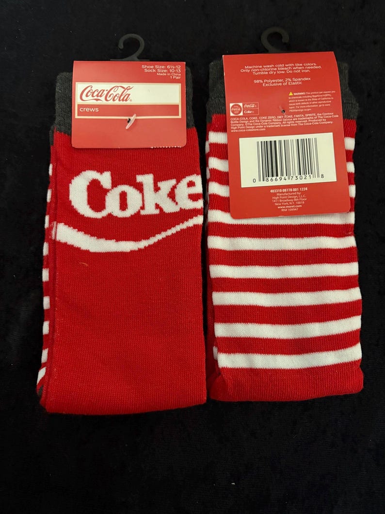 May include: Pair of red and white crew socks with the Coca-Cola logo. One sock features the word "Coke" in white, while the other has red and white stripes. The socks have a gray cuff and a red tag.