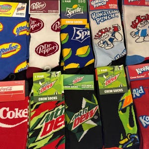 May include: Assortment of crew socks featuring various brand logos and designs. Includes socks with the logos of Yoohoo, Dr. Pepper, Sprite, Hawaiian Punch, Coca-Cola, Mountain Dew, and Kool-Aid. The socks are in various colors and patterns.
