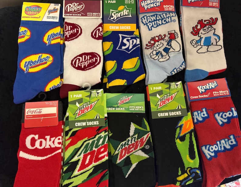 May include: Assortment of crew socks featuring various brand logos and designs. Includes socks with the logos of Yoohoo, Dr. Pepper, Sprite, Hawaiian Punch, Coca-Cola, Mountain Dew, and Kool-Aid. The socks are in various colors and patterns.