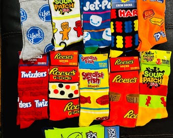 May include: A collection of 12 pairs of colourful crew socks featuring various candy brands, including Sour Patch Kids, Jet-Puffed, Haribo, Reese's Pieces, Swedish Fish, Twizzlers, and Jolly Rancher.
