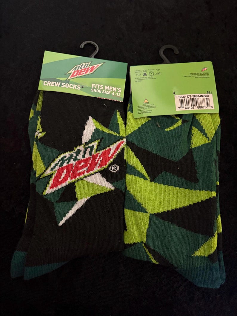 May include: Two pairs of crew socks featuring the Mountain Dew logo and geometric patterns in black, green, and white. The packaging reads "CREW SOCKS" and "FITS MEN'S SHOE SIZE 6-12".