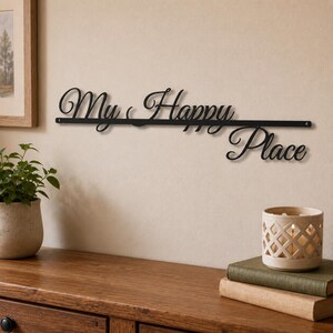 May include: A black metal wall sign featuring the cursive text "My Happy Place" mounted on a horizontal bar. The sign is constructed from thin, dark metal wire and includes small circular mounting holes at both ends of the straight base bar.