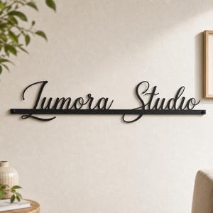 May include: A black metal wall sign featuring the text "Jumora Studio" in a cursive script font. The letters are connected to a thin, horizontal black metal bar that runs underneath the entire length of the word, secured by two small screws.