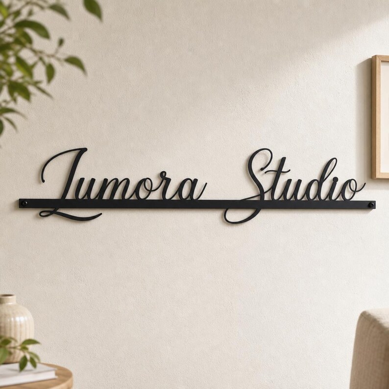 May include: A black metal wall sign featuring the text "Jumora Studio" in a cursive script font. The letters are connected to a thin, horizontal black metal bar that runs underneath the entire length of the word, secured by two small screws.