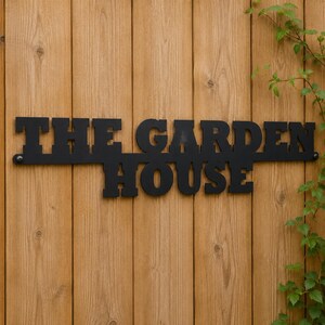 May include: A black metal sign featuring the words THE GARDEN HOUSE in a bold, block-style font. The sign is constructed as a single piece with mounting holes on the far left and right ends, displaying a matte finish and clean, sharp edges.