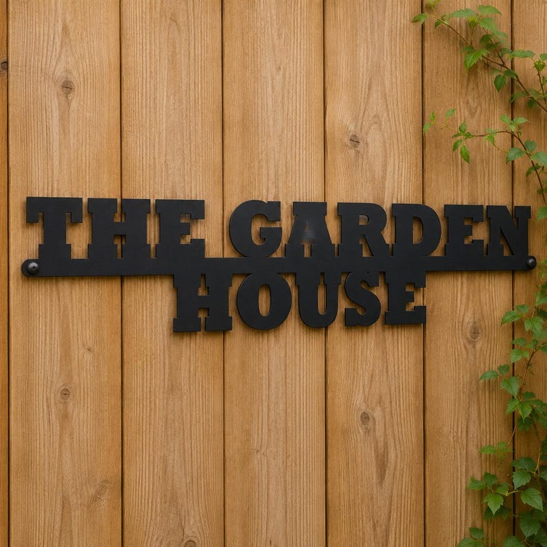 May include: A black metal sign featuring the words THE GARDEN HOUSE in a bold, block-style font. The sign is constructed as a single piece with mounting holes on the far left and right ends, displaying a matte finish and clean, sharp edges.