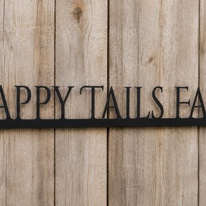 May include: A black metal sign featuring the words HAPPY TAILS FARM in a serif font. The letters are connected by a horizontal bar at the base, which includes a small circular mounting hole on each end for attachment to a flat surface.