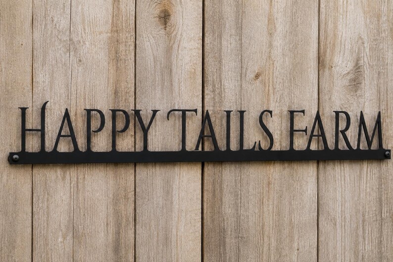 May include: A black metal sign featuring the words HAPPY TAILS FARM in a serif font. The letters are connected by a horizontal bar at the base, which includes a small circular mounting hole on each end for attachment to a flat surface.