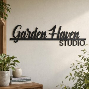 May include: A black metal wall sign featuring the words Garden Haven in a cursive script font, with the word STUDIO in a block sans-serif font positioned below on the right side, all mounted on a thin horizontal bar with visible screw holes.