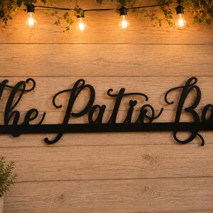 May include: A black metal wall sign featuring the cursive text The Patio Bar. The sign is constructed from a single piece of metal with a horizontal bar at the base and circular mounting holes on each end, displaying a clean, dark finish.