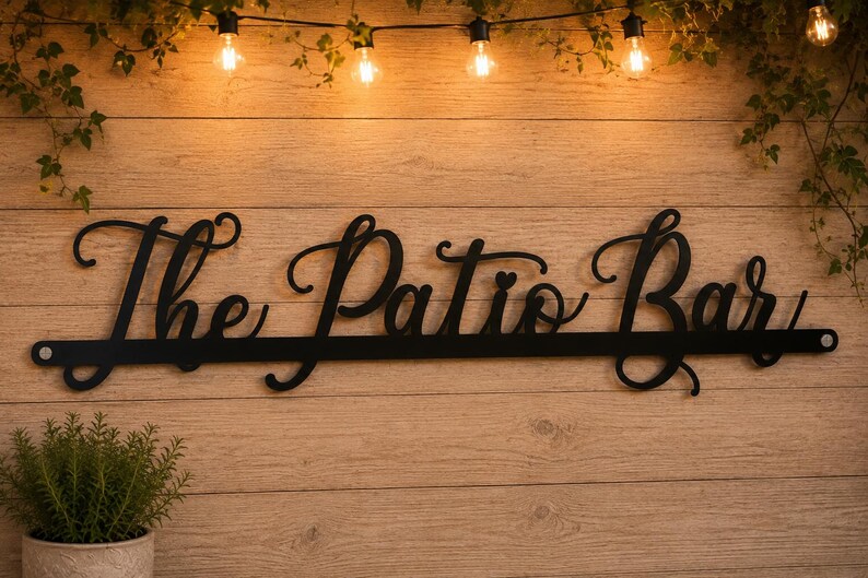 May include: A black metal wall sign featuring the cursive text The Patio Bar. The sign is constructed from a single piece of metal with a horizontal bar at the base and circular mounting holes on each end, displaying a clean, dark finish.