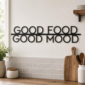May include: A black metal wall sign featuring the text GOOD FOOD GOOD MOOD in a sans-serif font. The words are arranged in two lines separated by a horizontal bar, with the letters connected to the bar. The sign is mounted on a plain wall.