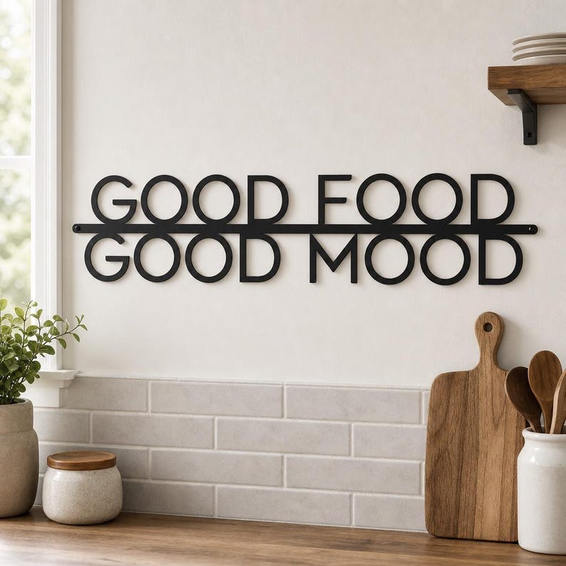 May include: A black metal wall sign featuring the text GOOD FOOD GOOD MOOD in a sans-serif font. The words are arranged in two lines separated by a horizontal bar, with the letters connected to the bar. The sign is mounted on a plain wall.