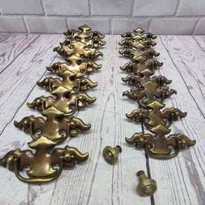 May include: A collection of brass-toned metal drawer pulls featuring ornate, symmetrical backplates with curved edges and swinging bail handles, accompanied by two small, circular matching knobs, all arranged in two vertical rows on a surface.