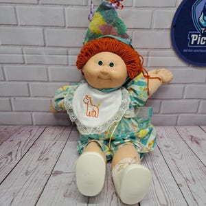 May include: A vintage Cabbage Patch Kid doll with red yarn hair, a white bib with a giraffe design, and a colorful hat. The doll is wearing a teal and yellow patterned outfit and white shoes.