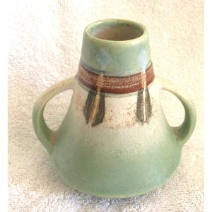 May include: A ceramic vase with two handles, featuring a tapered neck and a wide base. The exterior is finished in a matte light green glaze, accented by a horizontal brown band and vertical dark brown arrow-like motifs around the upper section.