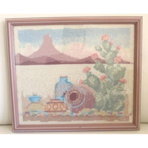 Può includere: Opera d'arte a punto croce incorniciata con un paesaggio desertico con una catena montuosa, cactus con fiori rosa e ceramiche. La tavolozza dei colori comprende sfumature di blu, rosa e marrone. La cornice è di colore marrone chiaro.