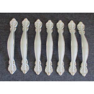 May include: Seven white cabinet pulls with gold trim are arranged on a gray surface. The pulls have a curved design with decorative detailing at each end. The overall aesthetic is vintage or antique.