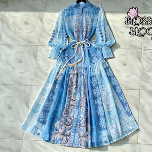 May include: A long, light blue dress with a high collar and long sleeves. The dress features a detailed pattern in shades of blue and white, with a matching belt. The brand name "Blossom Bloom" is visible in the upper right corner.