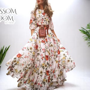 Könnte beinhalten: Ein weißes Maxikleid mit Blumenmuster mit roten, gelben und orangen Blumen. Das Kleid hat kurze Ärmel und einen braunen Gürtel mit einer kleinen braunen Tasche. Das Kleid ist von Blossom Bloom by Sbor.