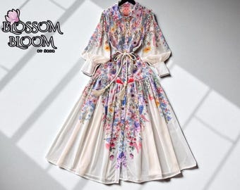Puede incluir: Un vestido largo con estampado floral con botones en la parte delantera, mangas largas y cinturón anudado. El vestido presenta un colorido estampado floral con tonos de rosa, morado, amarillo y azul sobre un fondo crema. La marca "Blossom Bloom" es visible.