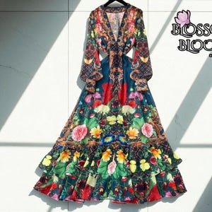 Peut inclure: Longue robe fluide avec un imprimé floral vibrant dans les tons de rouge, rose, jaune et vert. La robe présente des manches longues, un col en V et des découpes à la taille. Le logo Blossom Bloom est visible en haut à droite.