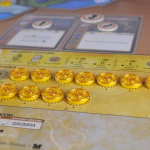 May include: A board game setup with yellow tokens and cards. The game board is tan with text and graphics. The tokens are circular with a yellow center and a gold rim. Two cards are in the background.