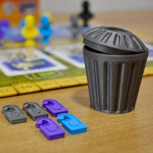 May include: A gray miniature trash can with a slightly open lid, next to several colored game pieces. The game pieces are gray, purple, and blue, shaped like door hangers. A board game is visible in the background.